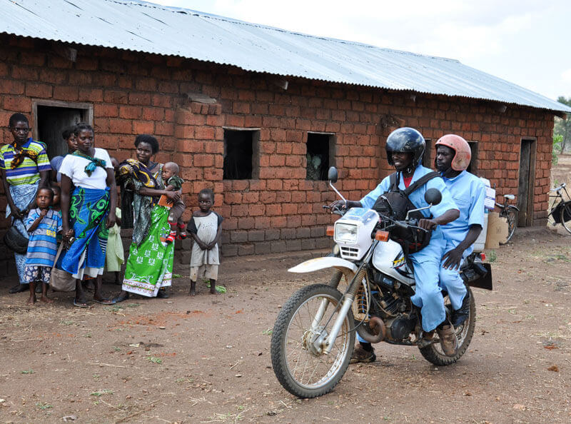Two male health workers on a motorcycle carrying a vaccine carrier and supplies, pulling up in front of a building where women and children are waiting. (PATH/Doune Porter)